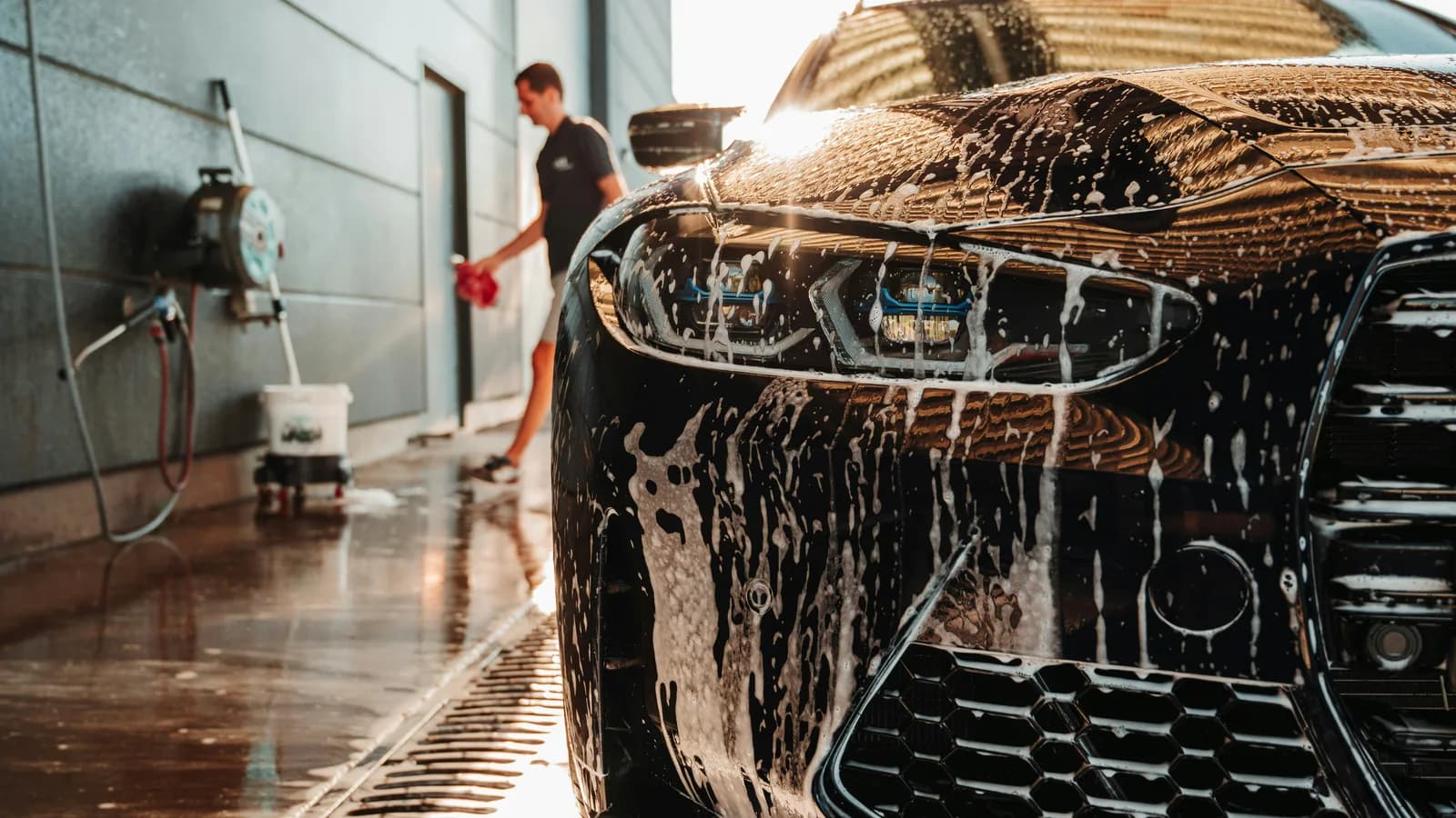 Photo by lucas clarysse on Unsplash A black car covered in soap foam sits in a wash bay while a detailer in the background holds a red towel beside a wall-mounted hose reel and a bucket cart.
