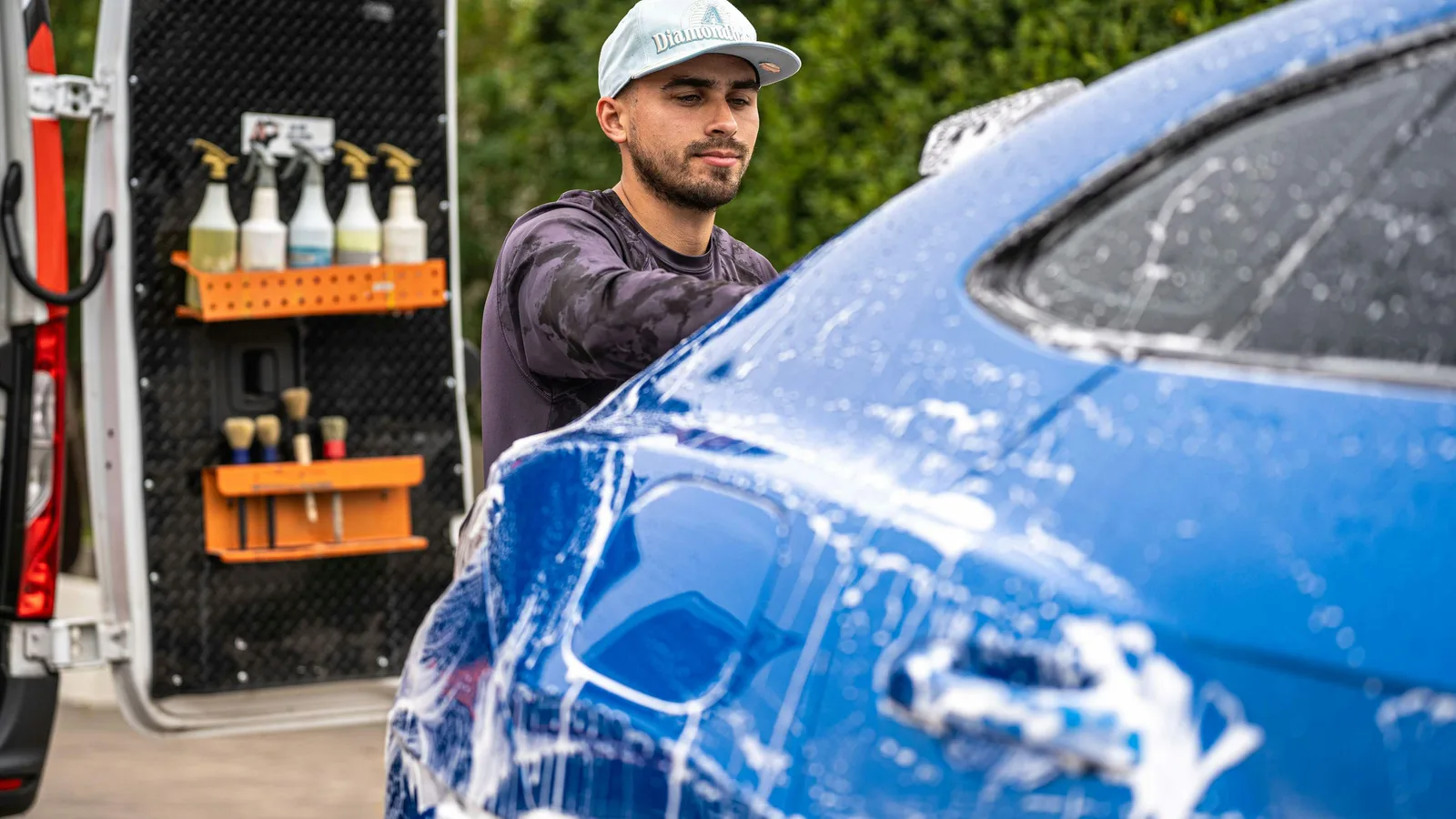 Photo by Daniel's Detailing on Unsplash A detailer foams a blue car beside an open work van with spray bottles and brushes racked on the door.