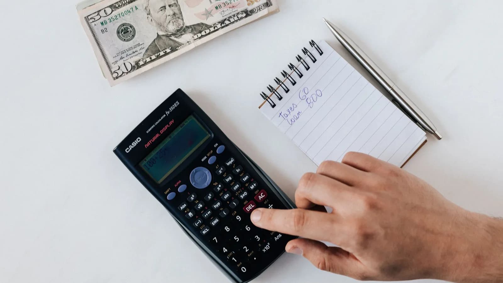 A hand using a calculator beside a notebook and dollar bills.